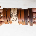 two hands holding an assortment of matching leather couples bracelets. The bracelets are engraved with personalized text like wifey, hubby, a third anniversary date, custom navigation coordinates.